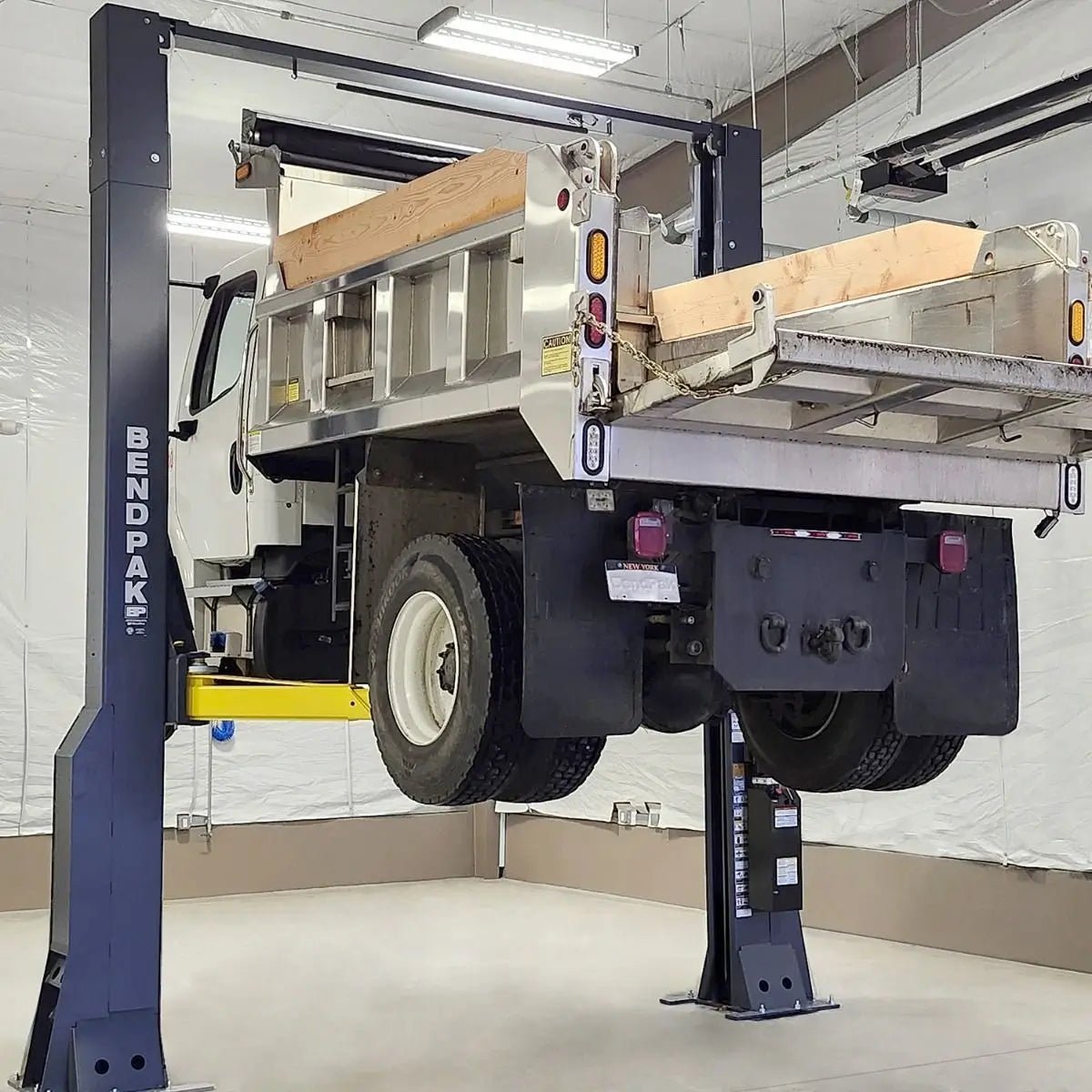 BendPak 20AP-192 two-post lift raising a dump truck in a fleet maintenance bay, showing yellow arms, gray columns, and clearfloor design suited for heavy vehicle service.