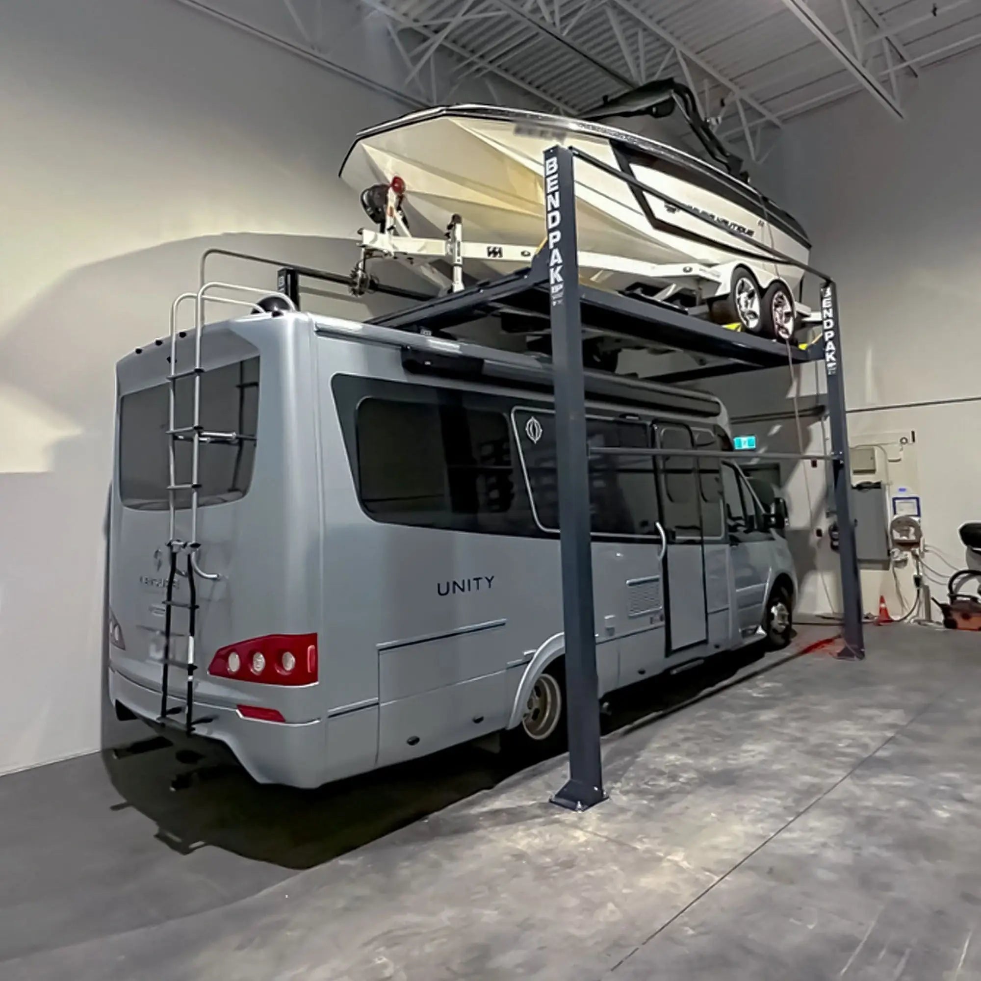 BendPak HD-7500PBX high-rise four-post boat and vehicle storage lift in warehouse use, holding a large white powerboat on the upper platform with a silver Unity RV parked below, showcasing tall steel columns, heavy-duty runways, and industrial-grade structure inside a modern facility.