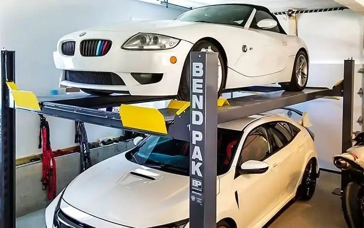 BendPak HD-9XL four-post lift installed in a home garage, holding two vehicles in stacked configuration with steel posts, safety locks, and yellow approach ramps for secure car storage.