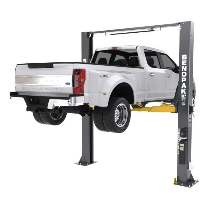 BendPak XPR-12CL two-post lift raising a Ford Super Duty dually; rear three-quarter view showing wide stance and adjustable lift arms for heavy-duty trucks.