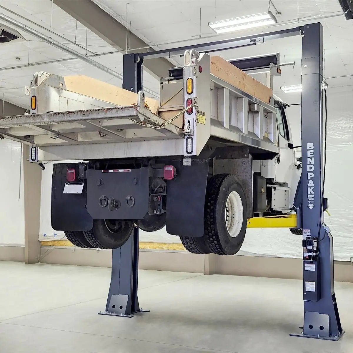 BendPak 20AP-192 two-post lift raising a large dump truck inside service bay, showing heavy-duty construction with yellow arms, tall gray columns, and overhead beam.