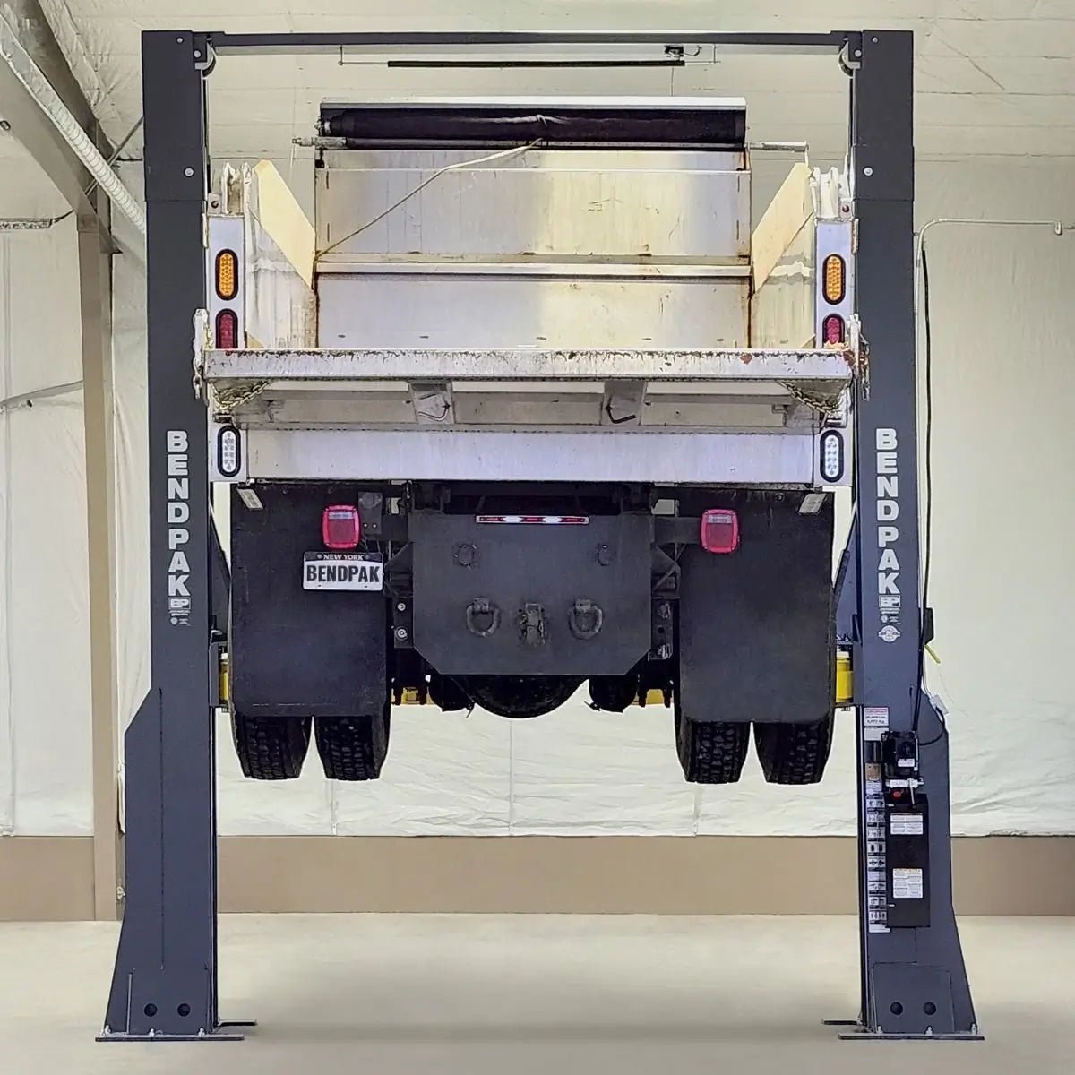 BendPak 20AP-192 two-post lift raising a dump truck viewed from the rear, showing tall steel columns, yellow lift arms, and overhead beam inside a service facility.