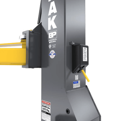 BendPak 20AP-192 two-post lift closeup of safety lock mechanism on column showing control lever, lock housing, warning labels, and yellow lift arm attachment point.