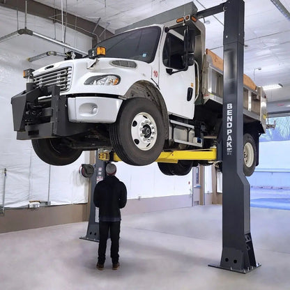 BendPak 20AP-192 two-post lift hoisting a heavy-duty dump truck, technician standing below, showcasing tall-rise extended height design and yellow lifting arms in service bay.