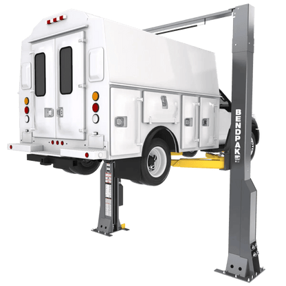 BendPak 20AP-192 two-post lift raising a white service truck, rear-left angle view showing extended-height clearfloor design, yellow lift arms, and gray steel columns with BendPak logo.