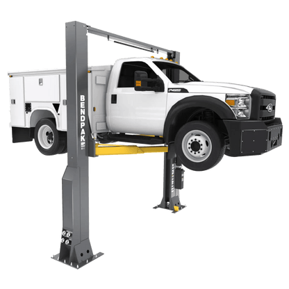 BendPak 20AP two-post lift raising a white service truck, angled front-left view showing heavy-duty construction, yellow arms, overhead beam, and BendPak-branded columns.