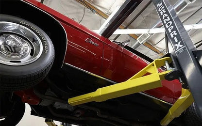 BendPak GP-7 two-post lift supporting a red classic car from below, showing yellow arms engaged under chassis in a low-ceiling garage installation.