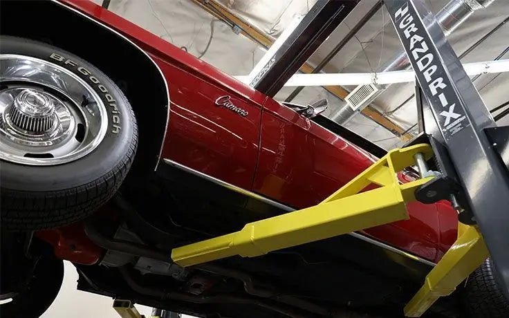 BendPak GP-7 two-post lift supporting a red classic car from below, showing yellow arms engaged under chassis in a low-ceiling garage installation.