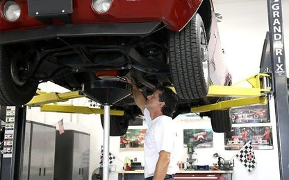 BendPak GP-7 two-post lift holding a red car overhead while a technician performs an oil drain service, showing undercarriage clearance and lift stability.