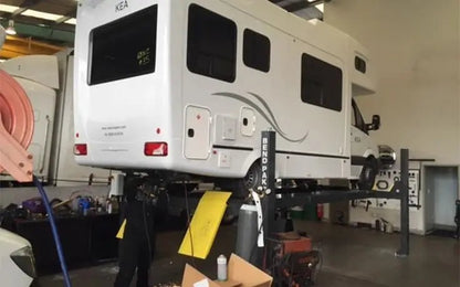 BendPak HD-14T tall four-post lift shown raising a large camper RV in a workshop, demonstrating extended post height, yellow ramps, and heavy-duty support for high-clearance vehicles.
