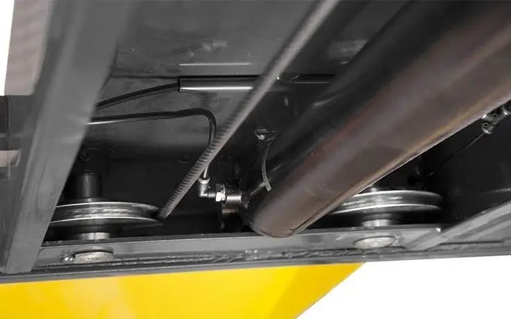 BendPak HD-9SW four-post lift underside view showing large cable sheave assembly with dual pulleys, cross shaft, and steel cables designed to reduce cable wear.