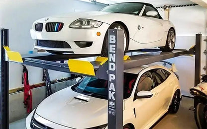 BendPak HD-9XL four-post lift installed in a home garage, holding two vehicles in stacked configuration with steel posts, safety locks, and yellow approach ramps for secure car storage.