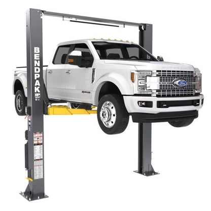 BendPak XPR-12CL two-post lift supporting a Ford Super Duty dually; front-left view highlighting heavy-duty arm reach and overhead hydraulic connection.