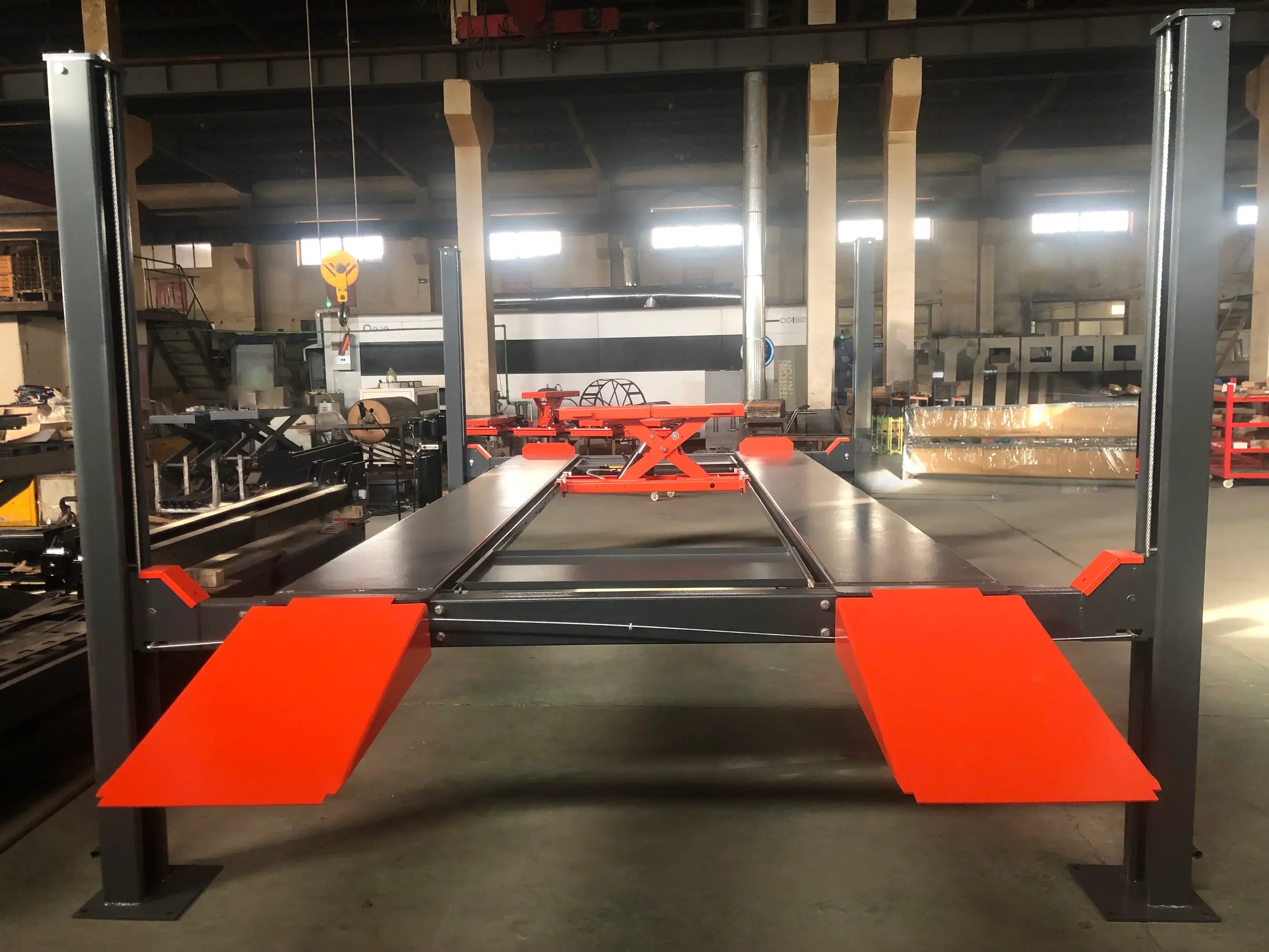 Katool KT-4H110 11,000 lb 4 post lift shown fully assembled in a warehouse, featuring a drive-on platform with orange safety ramps, hydraulic scissor jack in the center, and gray steel columns.