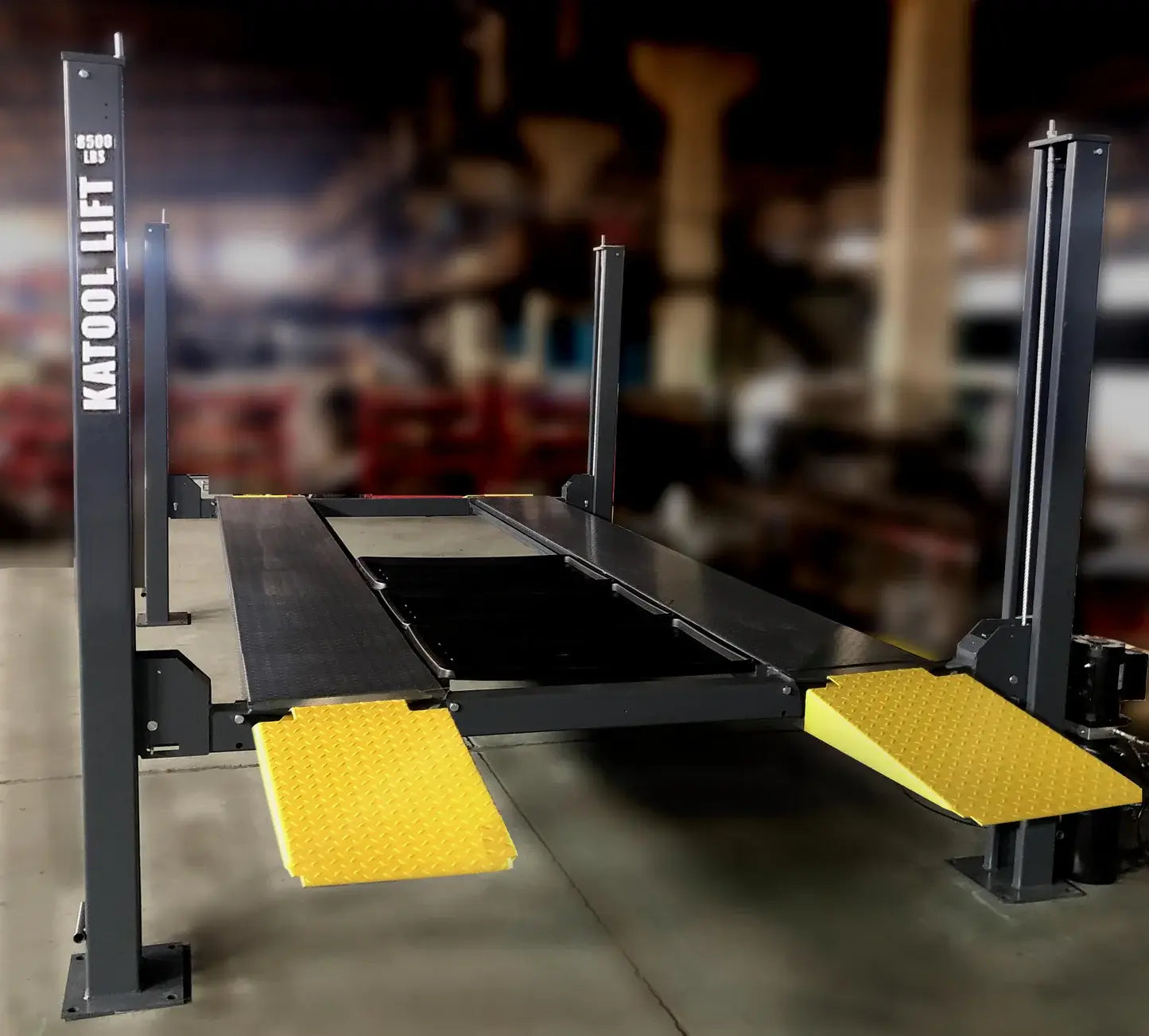 Katool KT-4H850 4-Post Car Lift in a garage, four posts, dual black runways, and yellow diamond-plate ramps with a blurred background.